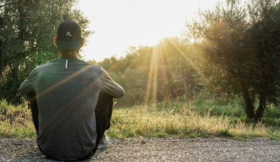 A person wearing a dark jacket and cap sits on a path, facing a grassy field and trees as sunlight filters through, creating bright rays in the serene outdoor setting.