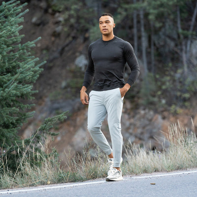 A man wearing a moisture-wicking black long-sleeve shirt, RHOBACK Delta Performance Jogger | Solid - Thunder Grey pants, and white sneakers walks along a road with grass, trees, and a rocky hillside in the background.
