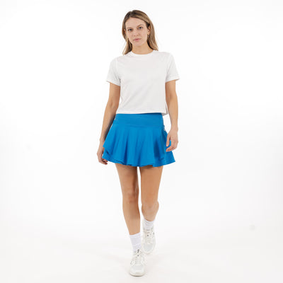 A woman stands against a white background wearing the RHOBACK Sydney Tennis Skort | Solid - Birdie Blue, featuring a high-rise ribbed waistband. She pairs it with a white t-shirt, socks, and sneakers, gazing forward with a neutral expression.