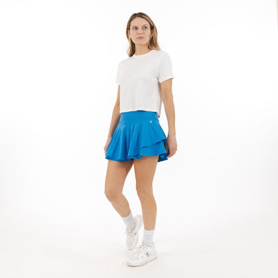 A woman stands against a plain white background wearing the RHOBACK Sydney Tennis Skort | Solid - Birdie Blue, featuring a bright blue ruffled design and high-rise ribbed waistband, paired with a white t-shirt, white socks, and sneakers.