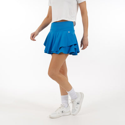 A person wears a white cropped t-shirt and the RHOBACK Sydney Tennis Skort | Solid - Birdie Blue with a high-rise ribbed waistband, paired with white crew socks and sneakers, standing against a plain white background. Their face is not visible.