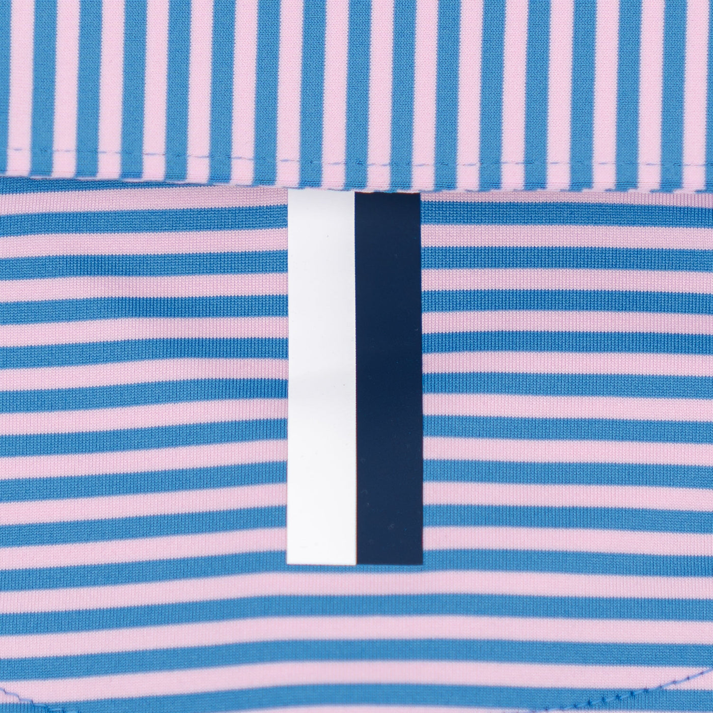 Close-up of RHOBACK’s The Birdie Stripe Performance Polo in Birdie Blue/Lotus Pink, showing four-way stretch fabric with blue and pink stripes, central vertical tag, and bottom seam for comfort and easy movement.