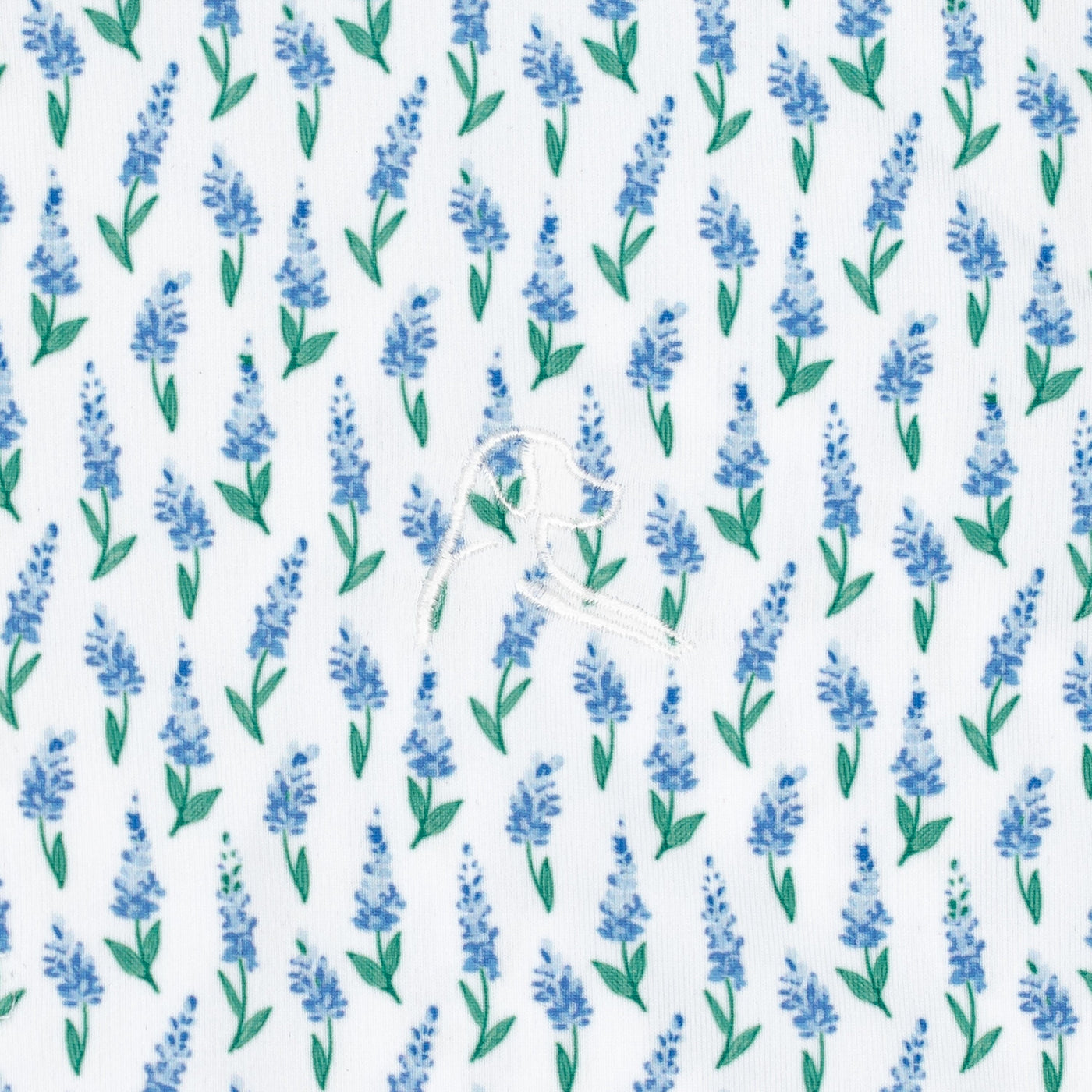 The Lil' Blue Bonnet 2.0 Performance Polo by RHOBACK features a playful white fabric with blue lavender sprigs, green leaves, and a stitched dog outline—delivering sun protection and charm for toddlers.