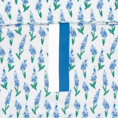 A close-up of RHOBACK's The Lil' Blue Bonnet 2.0 Performance Polo in White/Lake Blue, featuring a repeating blue bonnet floral and green leaf pattern on white fabric, with a vertical white and blue stripe down the center.