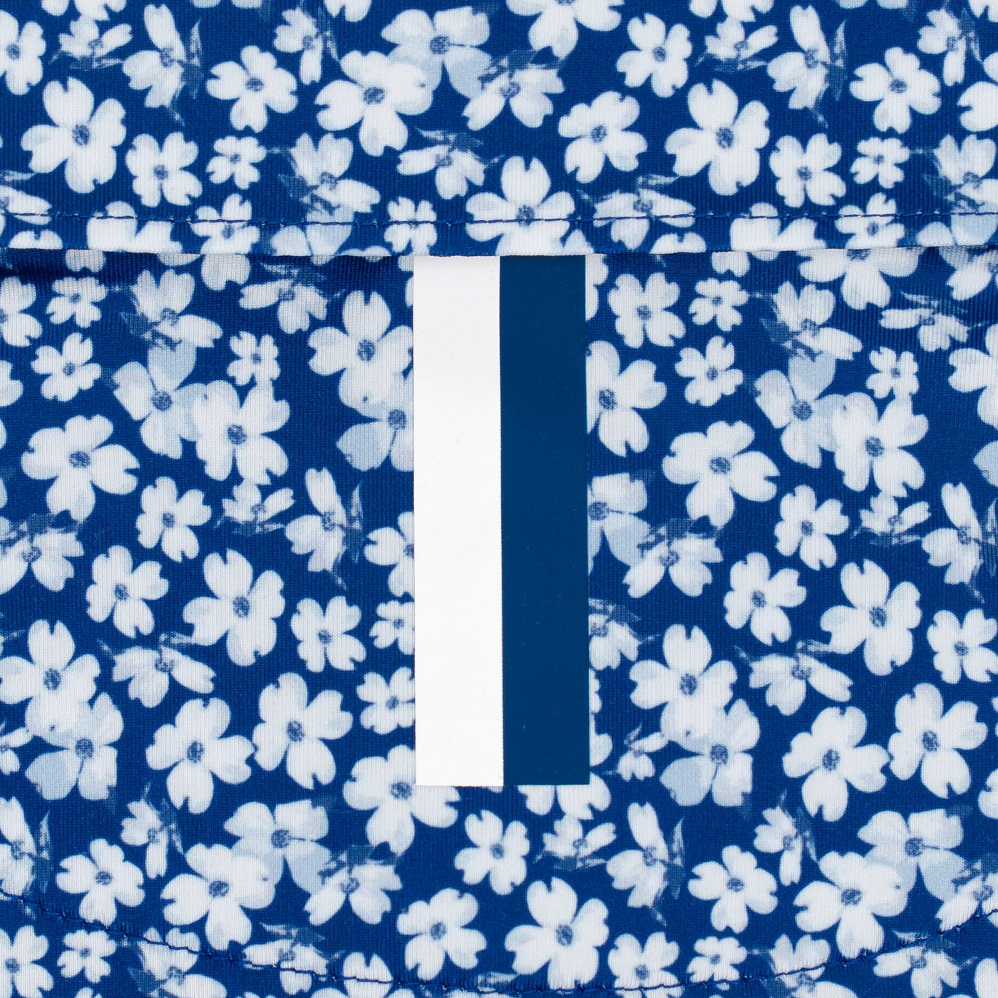 Close-up of blue fabric with a white and light blue floral pattern, featuring moisture-wicking material and a white label with a navy stripe—ideal for RHOBACK’s The Dogwood 2.0 Performance Polo in Shoal Blue/White.