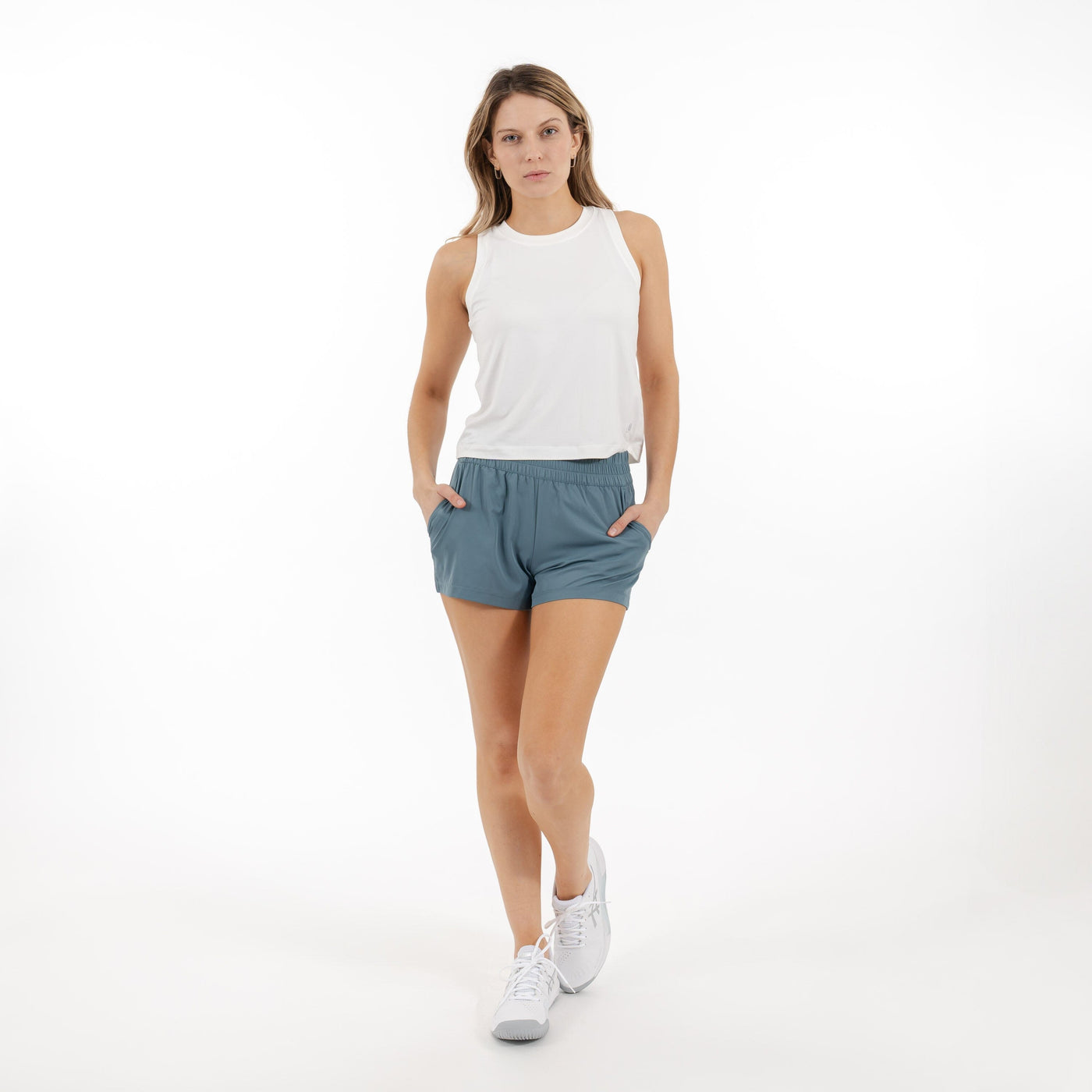 A woman wearing a white sleeveless top, RHOBACK Era High-Rise Short | Solid in Jungle Green, and white sneakers stands with her hands in her pockets against a plain white background.