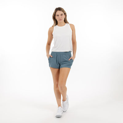 A woman wearing a white sleeveless top, RHOBACK Era High-Rise Short | Solid in Jungle Green, and white sneakers stands with her hands in her pockets against a plain white background.