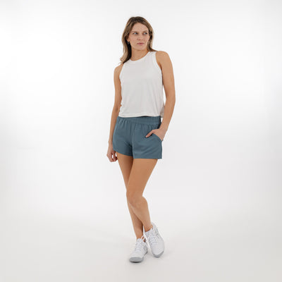 A woman wears a sleeveless white top, RHOBACK Era High-Rise Short in Jungle Green, and white sneakers, standing on a white background with one hand in her pocket, looking off to the side.