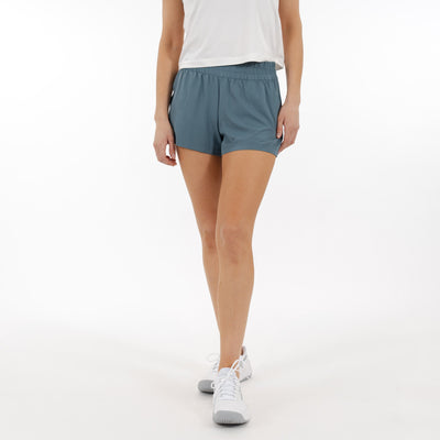 A person wears a white sleeveless top, RHOBACK Era High-Rise Short | Solid - Jungle Green made from four-way stretch, moisture-wicking fabric, and white sneakers. Standing against a white background with head and lower arms out of frame.