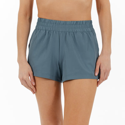A person wears RHOBACK's Era High-Rise Short | Solid in Jungle Green, featuring a four-way stretch and elastic waistband, standing against a white background with their upper body and most of their legs visible.