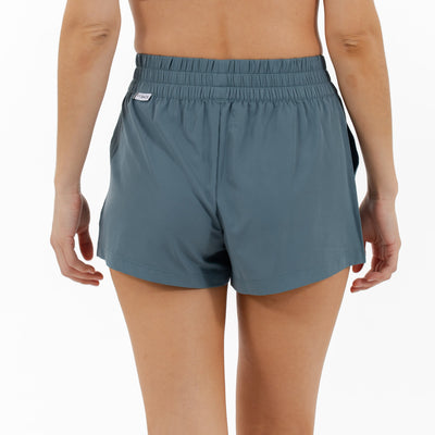 A person faces away from the camera, wearing RHOBACK's Era High-Rise Short | Solid in Jungle Green, featuring four-way stretch fabric and an elastic waistband, set against a plain white background.