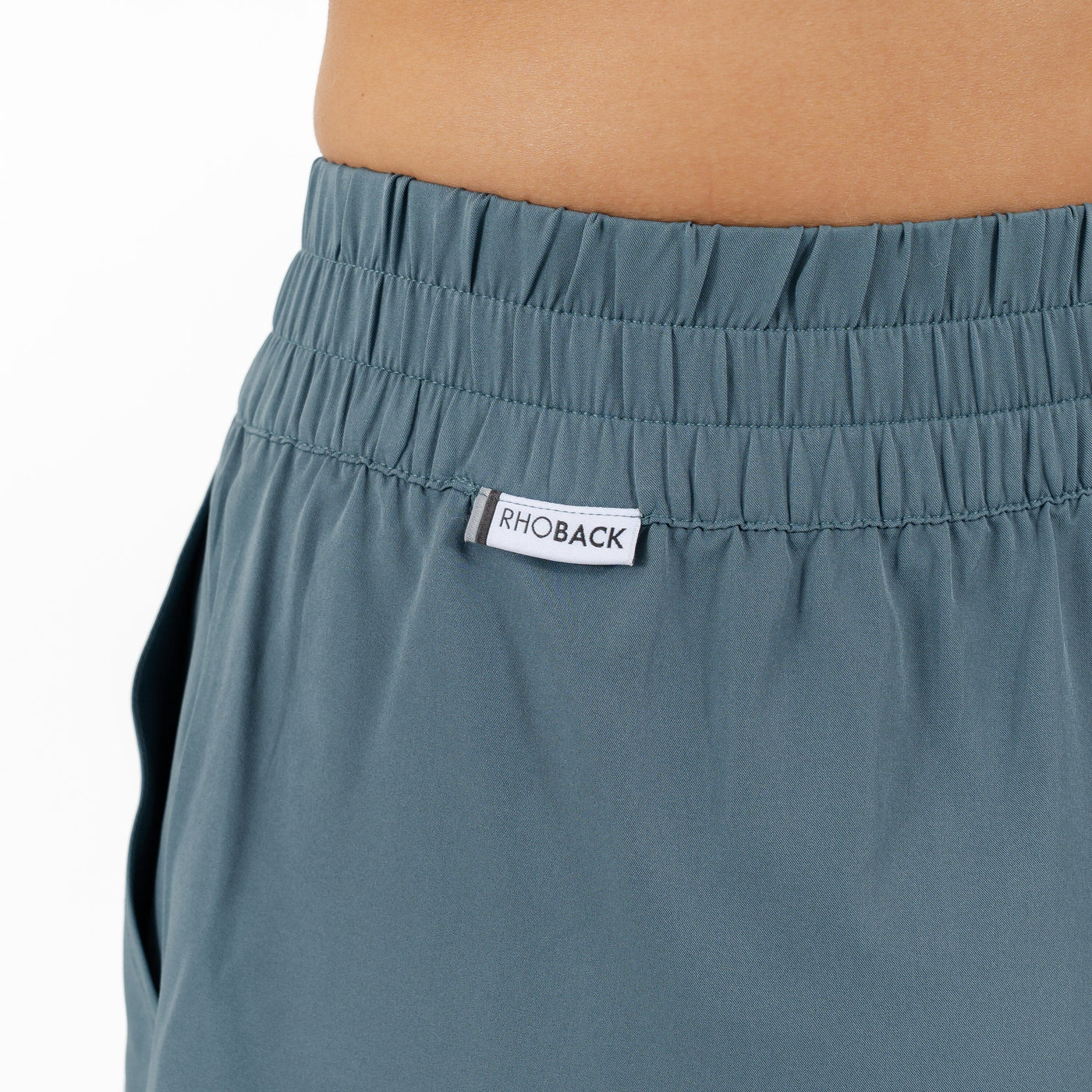 Close-up of a person wearing the RHOBACK Era High-Rise Short in Solid Jungle Green. These shorts feature an elastic waistband, a "RHOBACK" label at the front, moisture-wicking fabric, and a left-side pocket.