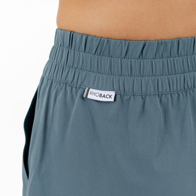Close-up of a person wearing the RHOBACK Era High-Rise Short in Solid Jungle Green. These shorts feature an elastic waistband, a "RHOBACK" label at the front, moisture-wicking fabric, and a left-side pocket.