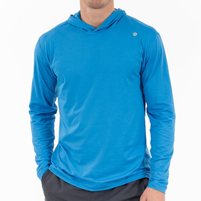 A man wearing the RHOBACK Blitz Tech Hoodie in Heather - Birdie Blue/Sapphire Blue stands with his hands in his pockets against a plain white background, his face partially out of frame.