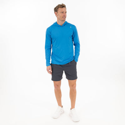 A man stands against a white background wearing the RHOBACK Blitz Tech Hoodie in Birdie Blue/Sapphire Blue, made with moisture-wicking SilverKnit fabric, paired with dark gray shorts and white athletic shoes. He looks left with one hand in his pocket.
