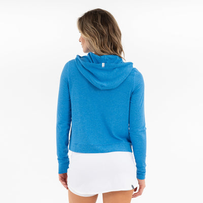 A woman with wavy, shoulder-length hair is seen from behind, wearing an RHOBACK Tempo Performance Hoodie in Birdie Blue/Birdie Blue Heather and a white skirt, standing against a plain white background.