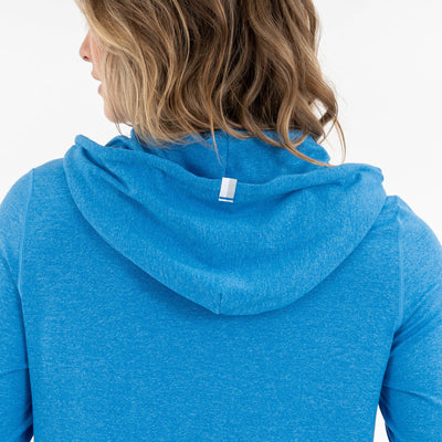 A person with wavy, light brown hair is seen from behind, wearing a RHOBACK Tempo Performance Hoodie in Birdie Blue. The hood is up but not on the head, and a small white tag accents the moisture-wicking VSoft™ fabric.