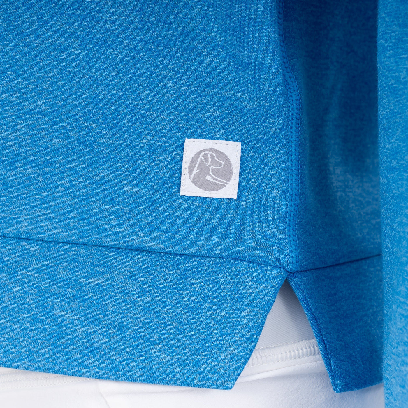 A close-up of the RHOBACK Tempo Performance Hoodie in Heather - Birdie Blue/Birdie Blue, made from VSoft™ fabric, shows a small white tag with a flexed arm icon, worn over white pants.