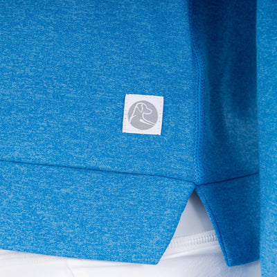 A close-up of the RHOBACK Tempo Performance Hoodie in Heather - Birdie Blue/Birdie Blue, made from VSoft™ fabric, shows a small white tag with a flexed arm icon, worn over white pants.