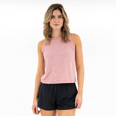 A woman with wavy, shoulder-length hair wears the RHOBACK Drift Performance Crop Tank in Heather - Dusk Pink/Lotus Pink and black shorts, standing against a plain white background.
