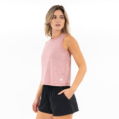 A woman wears the RHOBACK Drift Performance Crop Tank in Heather - Dusk Pink/Lotus Pink, featuring four-way stretch and moisture-wicking fabric, as she stands with one hand in her pocket against a plain white background.