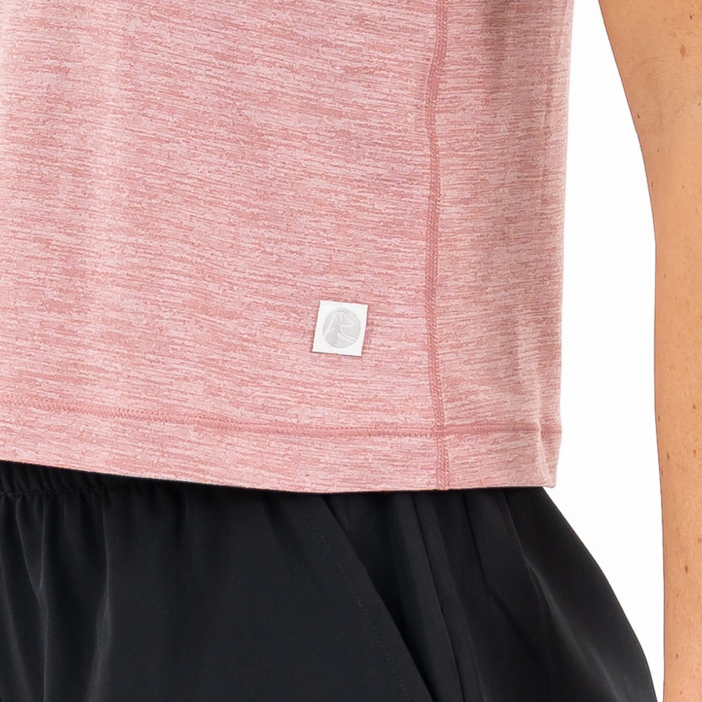 A close-up of a person wearing the RHOBACK Drift Performance Crop Tank in Heather - Dusk Pink/Lotus Pink, featuring SkyKnit™ fabric and a small white square logo near the hem, paired with black pants.