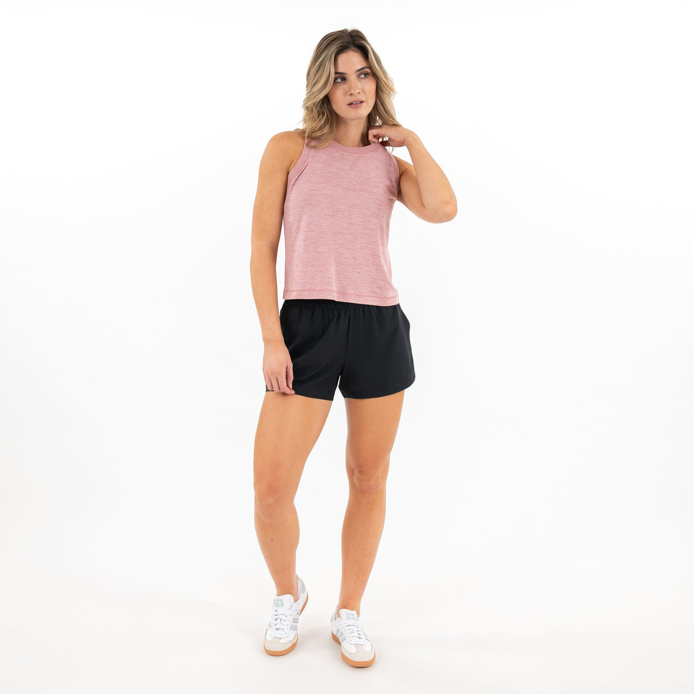 A woman with light brown hair stands against a white background wearing the RHOBACK Drift Performance Crop Tank in Heather Dusk Pink/Lotus Pink, paired with black shorts and white sneakers, looking to the side with one hand touching her neck.