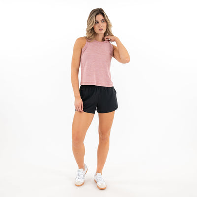 A woman with light brown hair stands against a white background wearing the RHOBACK Drift Performance Crop Tank in Heather Dusk Pink/Lotus Pink, paired with black shorts and white sneakers, looking to the side with one hand touching her neck.