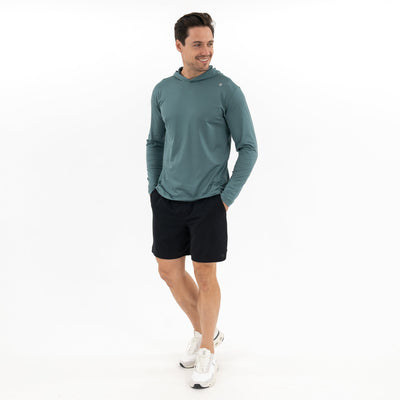 A man wearing the RHOBACK Blitz Tech Hoodie | Heather - Jungle Green/Dusty Pine, black shorts, and white sneakers stands against a white background, smiling to the side with his hands in his pockets.