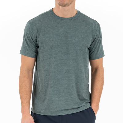 A person wearing the RHOBACK Tailwind Performance Tee in Heather - Jungle Green/Dusty Pine and dark pants stands with one hand in their pocket against a plain white background.
