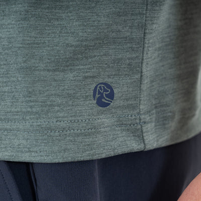 Close-up of the RHOBACK Tailwind Performance Tee in Heather - Jungle Green/Dusty Pine, made from four-way stretch SkyKnit™ fabric, featuring a small dark circular logo with an abstract dog’s head near the hem above dark pants.