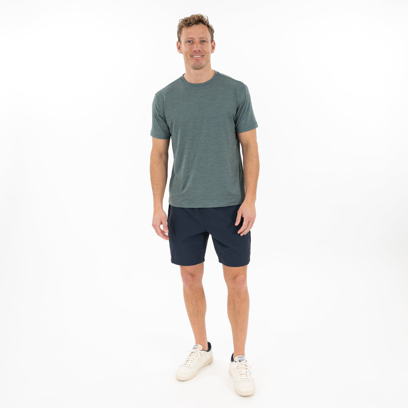 A man stands smiling against a plain white background, wearing the RHOBACK Tailwind Performance Tee in Heather - Jungle Green/Dusty Pine, paired with dark shorts and white sneakers.