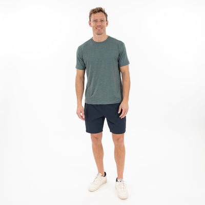 A man stands smiling against a plain white background, wearing the RHOBACK Tailwind Performance Tee in Heather - Jungle Green/Dusty Pine, paired with dark shorts and white sneakers.