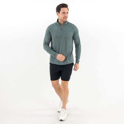 A man walks forward wearing the RHOBACK Tailwind Performance Q-Zip in Heather - Jungle Green/Dusty Pine, paired with black shorts and white sneakers against a plain white background.