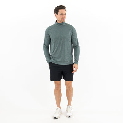 A man stands against a white background wearing the RHOBACK Tailwind Performance Q-Zip in Heather - Jungle Green/Dusty Pine, black shorts, and white sneakers. He looks to the side, right hand near his pocket.