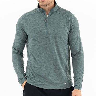 A man wearing the RHOBACK Tailwind Performance Q-Zip in Heather - Jungle Green/Dusty Pine with black pants poses with one hand in his pocket against a plain white background.