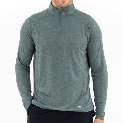 A man wears RHOBACK's Tailwind Performance Q-Zip in Heather - Jungle Green/Dusty Pine with black pants, standing against a plain white background. His face is partially out of frame.