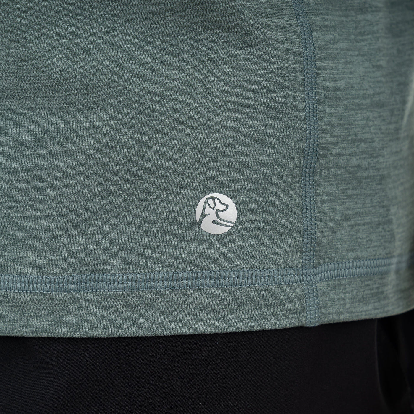 Close-up of the RHOBACK Tailwind Performance Q-Zip in Heather - Jungle Green/Dusty Pine, featuring visible stitching and a small circular dog head logo near the hem. The moisture-wicking shirt is shown above black pants.