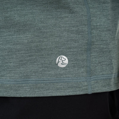 Close-up of the RHOBACK Tailwind Performance Q-Zip in Heather - Jungle Green/Dusty Pine, featuring visible stitching and a small circular dog head logo near the hem. The moisture-wicking shirt is shown above black pants.