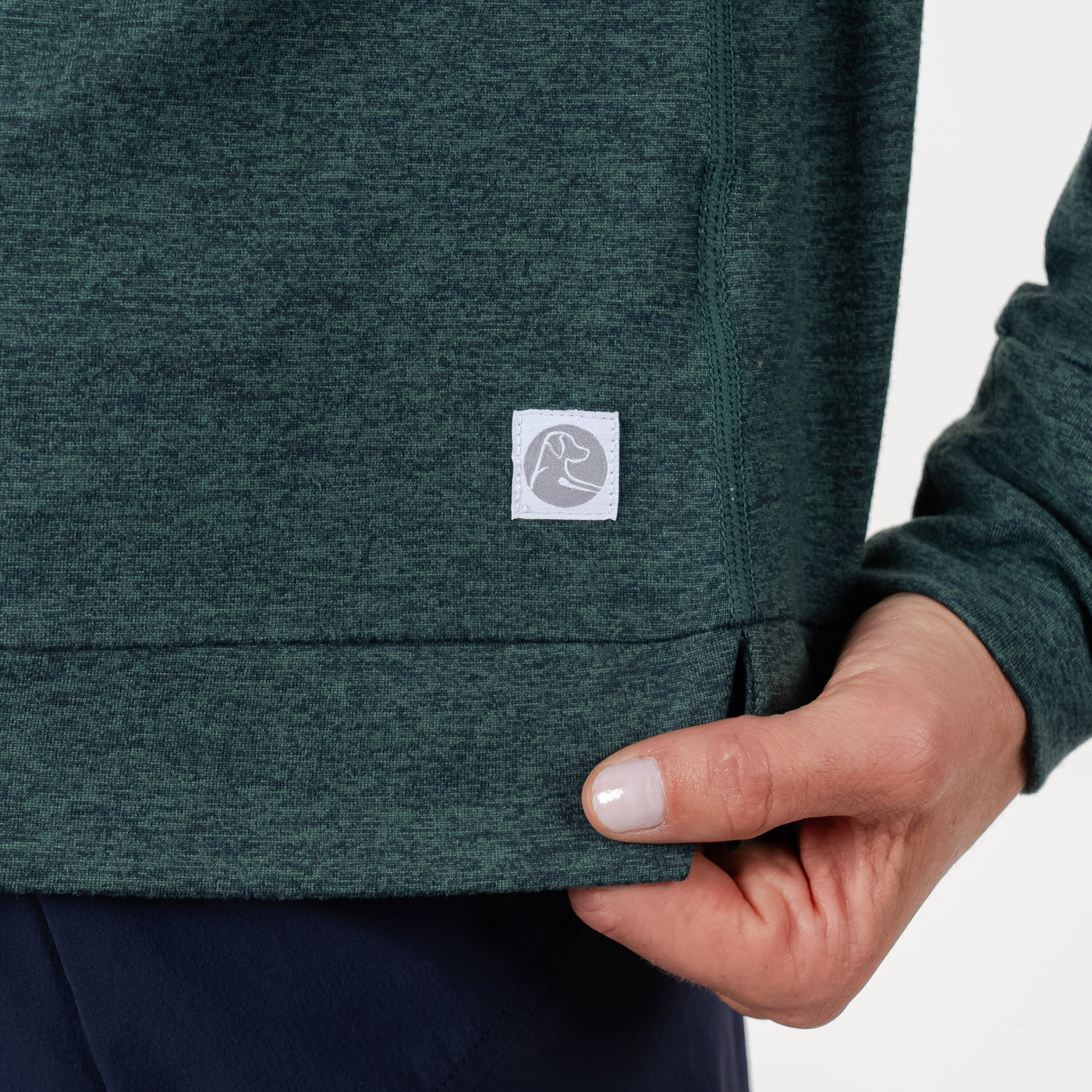 A close-up of a hand gently holding the bottom hem of the RHOBACK Tempo Performance Full-Zip Hoodie in Heather - Ponderosa Green/Willow Green, made from VSoft fabric with a small square white logo. The person is wearing dark pants.