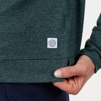 A close-up of a hand gently holding the bottom hem of the RHOBACK Tempo Performance Full-Zip Hoodie in Heather - Ponderosa Green/Willow Green, made from VSoft fabric with a small square white logo. The person is wearing dark pants.