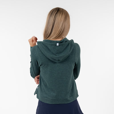 A woman with long blonde hair is seen from the back wearing the RHOBACK Tempo Performance Full-Zip Hoodie in Heather - Ponderosa Green/Willow Green, paired with a dark skirt, against a plain white background.