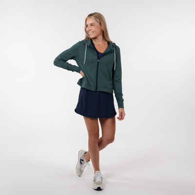 A woman smiles and looks to the side, wearing a RHOBACK Tempo Performance Full-Zip Hoodie in Heather - Ponderosa Green/Willow Green with moisture-wicking VSoft fabric over a navy dress and white sneakers, one hand on her hip and legs crossed.