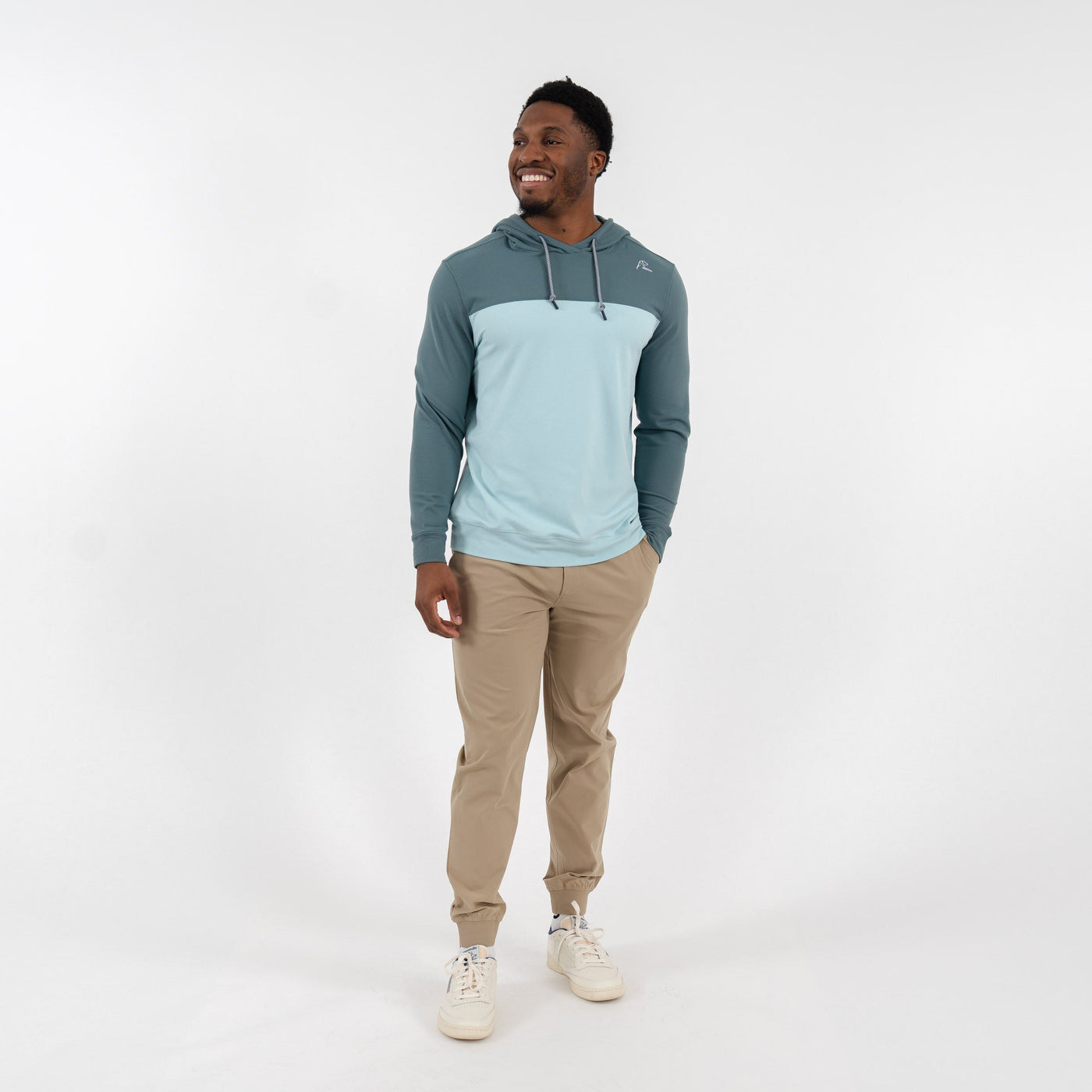A man wears the RHOBACK Hesi Performance Hoodie | Colorblock in Jungle Green/Aquafrost, beige pants, and white sneakers, smiling against a plain white background.