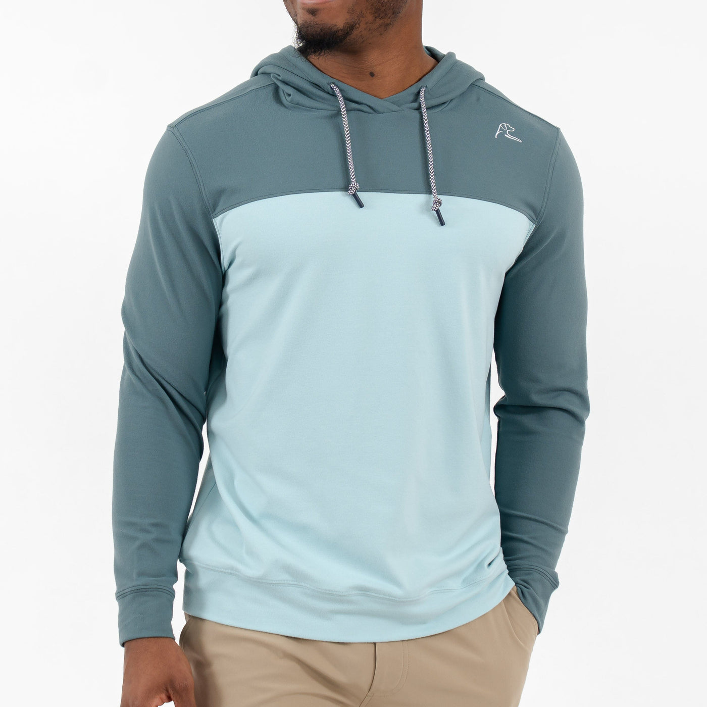 A man wears the RHOBACK Hesi Performance Hoodie | Colorblock - Jungle Green/Aquafrost, featuring teal sleeves and hood with a light blue torso, crafted from VSoft™ fabric, along with khaki pants, standing against a white background.