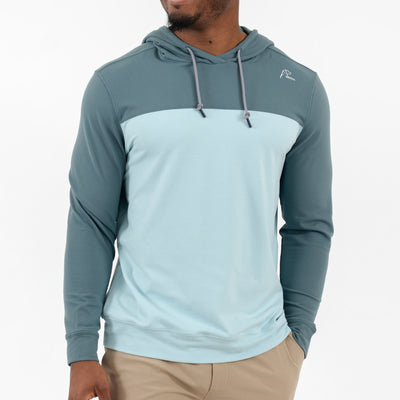 A man wears the RHOBACK Hesi Performance Hoodie | Colorblock - Jungle Green/Aquafrost, featuring teal sleeves and hood with a light blue torso, crafted from VSoft™ fabric, along with khaki pants, standing against a white background.