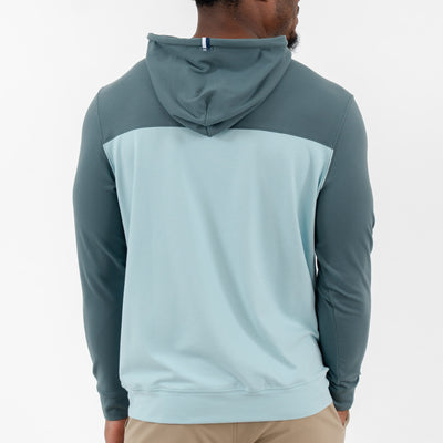 A person is seen from the back in a RHOBACK Hesi Performance Hoodie | Colorblock - Jungle Green/Aquafrost made with VSoft™ fabric, paired with beige pants. The hood is up against a plain white background.