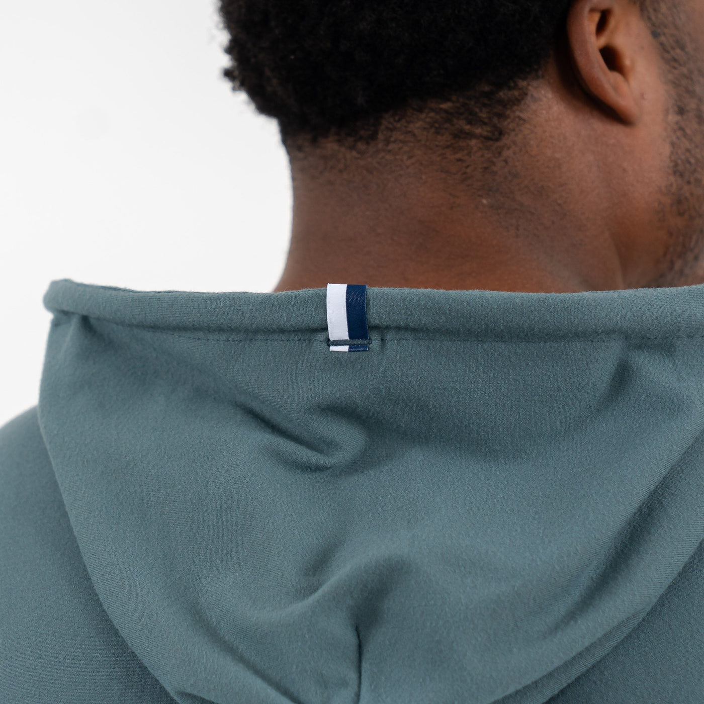 Close-up of the back of a person wearing the RHOBACK Hesi Performance Hoodie | Colorblock in Jungle Green/Aquafrost, made from VSoft™ fabric with a blue-and-white strip tag at the center of the hood. Face not visible.