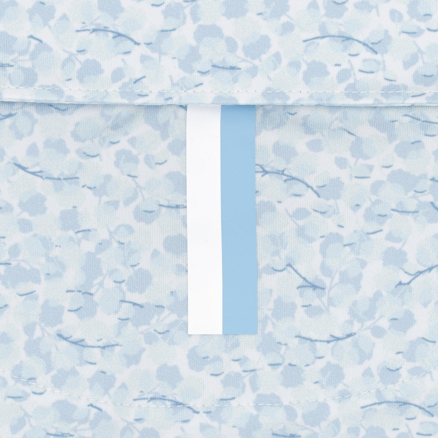 A close-up of the RHOBACK The Iridescence Performance Polo in White/Pure Blue, featuring breathable fabric with a light blue and white floral pattern and a vertical blue and white stripe down the center. Delicate abstract flowers in the background.