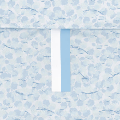 A close-up of the RHOBACK The Iridescence Performance Polo in White/Pure Blue, featuring breathable fabric with a light blue and white floral pattern and a vertical blue and white stripe down the center. Delicate abstract flowers in the background.